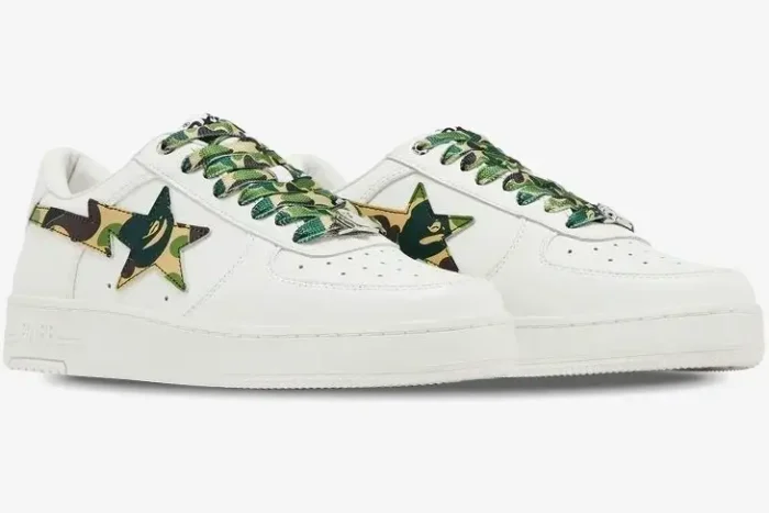 A pair of white Bape Sta Low sneakers with green camo star logos and laces, shown from a side angle. - SKU 1K70191335-GRN